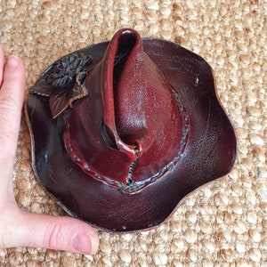 May include: A small, dark brown leather hat with a pointed top and a decorative flower design. The hat has a unique shape and is made of high-quality leather.