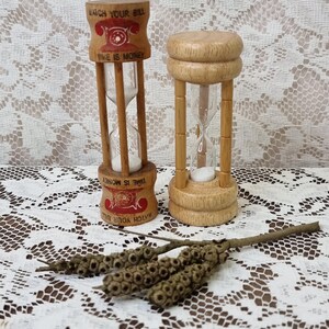 May include: Two wooden hourglasses with white sand. The left hourglass features a red telephone graphic and the text WATCH YOUR BILL and TIME IS MONEY. The right hourglass is plain light wood. A small brown twig with seed pods lies in front.