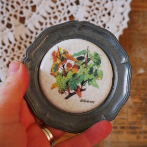 May include: A silver-colored metal coaster with a white ceramic tile insert featuring a painted design of grapes and leaves. The tile has the word "Silvaner" printed on it.