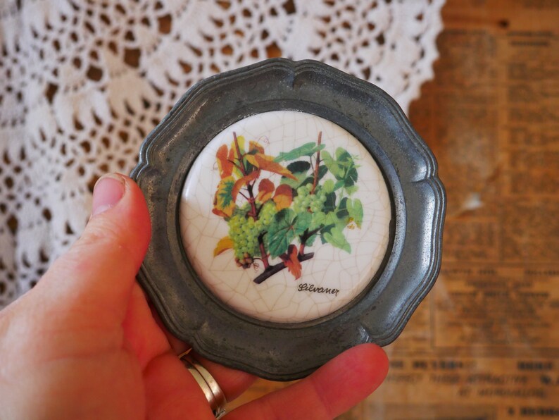 May include: A silver-colored metal coaster with a white ceramic tile insert featuring a painted design of grapes and leaves. The tile has the word "Silvaner" printed on it.
