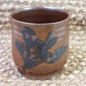 Könnte beinhalten: Eine kleine, braune Keramiktasse mit einem schwarzen Blumendesign. Die Tasse hat einen abgerundeten Boden und einen leicht ausgestellten Rand.