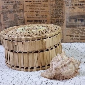 Op de afbeelding: Een geweven rieten mand met een deksel, versierd met witte en beige kralen. De mand staat op een wit kantkleedje naast een grote, beige schelp.