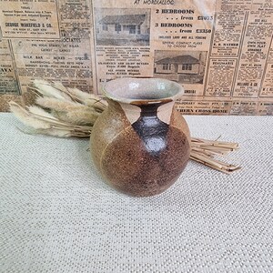 Peut inclure: Un vase en céramique brun et noir avec un bord brun clair. Le vase est posé sur une surface texturée brun clair.