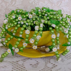 May include: A yellow teacup and saucer are filled with green beaded strands, decorated with white daisy accents and faux leaves. A silver spoon rests on the saucer, creating a whimsical floral display. A decorative floral arrangement.
