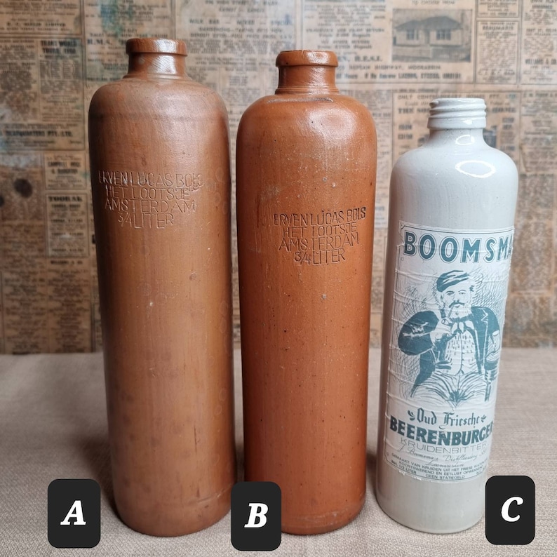 K&ouml;nnte beinhalten: Drei antike Glasflaschen. Zwei braune Flaschen mit gepr&auml;gtem Text, der "Erven Lucas Bois Het Lootse Amsterdam 3/4 Liter" lautet. Eine wei&szlig;e Flasche mit einem blauen und gr&uuml;nen Etikett, auf dem "Boomsma Oud Friesche Beerenburger" steht.
