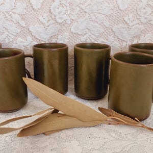 May include: A set of six cylindrical olive green ceramic mugs with dark brown handles. Each mug features a smooth, matte finish and a simple, straight-sided design. A few dried, light brown leaves are positioned in the foreground.