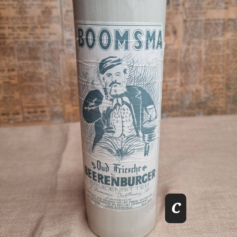 K&ouml;nnte beinhalten: Eine Vintage-Glasflasche mit einem Etikett, auf dem "Oud Friesche Beerenburger Kruidenbitter Bomma's Distilleerderij" steht. Das Etikett zeigt eine Comic-Illustration eines Mannes mit Bart und Pfeife, der ein Glas h&auml;lt. Die Flasche ist mit dem Markennamen "BOOMSMA" gekennzeichnet.