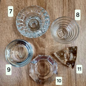 May include: An overhead shot displays five clear glass candle holders. The holders vary in shape, including round, hexagonal, and triangular designs. The glass has a reflective quality, and the holders are arranged on a wooden surface.