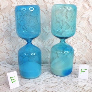 May include: Two blue hourglasses with a lace pattern on the top section. The hourglasses have a rounded base and top, connected by a narrow neck. The sand inside is a lighter shade of blue. The background is a white lace fabric.