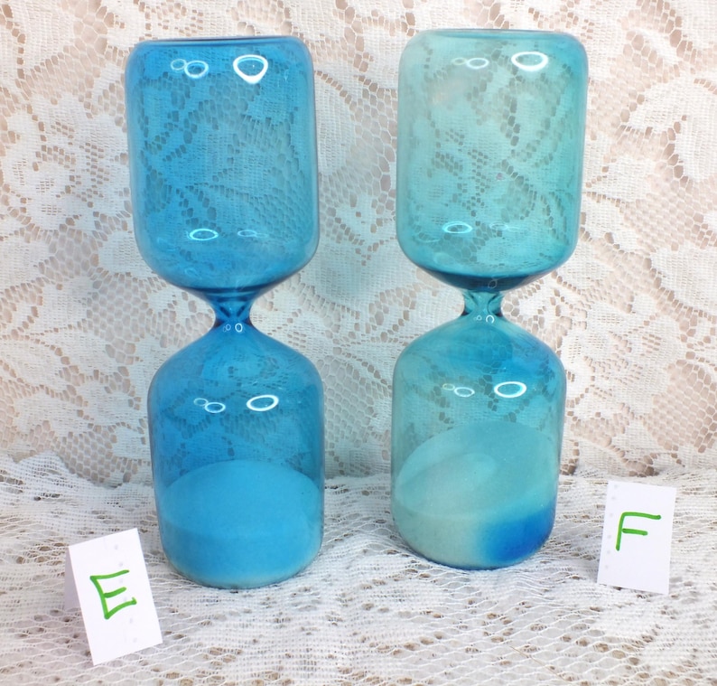 May include: Two blue hourglasses with a lace pattern on the top section. The hourglasses have a rounded base and top, connected by a narrow neck. The sand inside is a lighter shade of blue. The background is a white lace fabric.