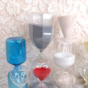 May include: A selection of glass hourglasses in different shapes and colours. Some contain red, white, and grey sand. Others are blue. The background is a white lace pattern.