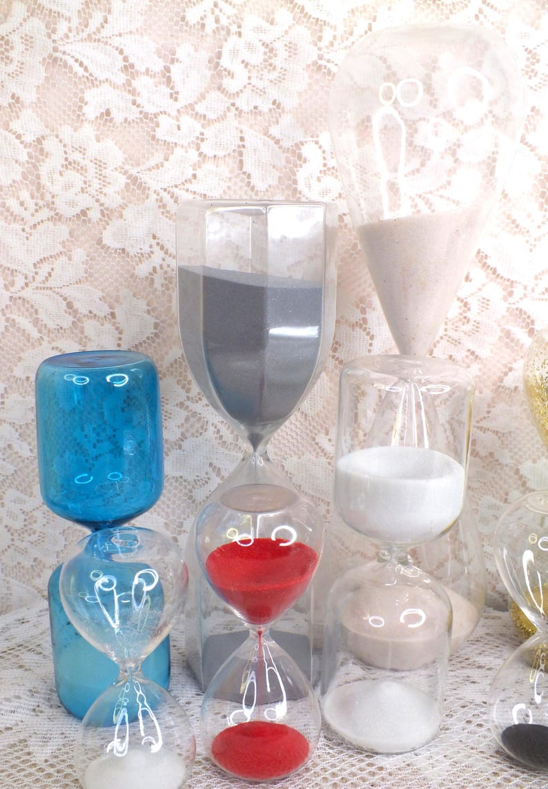 May include: A selection of glass hourglasses in different shapes and colours. Some contain red, white, and grey sand. Others are blue. The background is a white lace pattern.