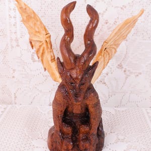 May include: A hand-carved wooden figurine of a seated creature with two long, spiralling horns and large, light-coloured wings extending outwards. The body of the figure is a dark brown, textured wood, while the wings are a lighter, natural wood tone.