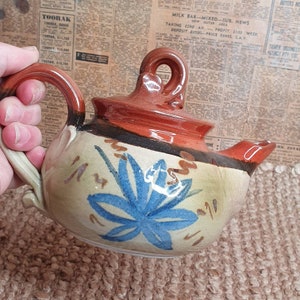 Può includere: Una teiera in ceramica dipinta a mano con un motivo floreale blu. La teiera ha un coperchio marrone e un manico marrone.