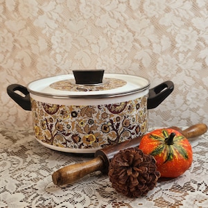 May include: A round white enamel pot with a brown, yellow, and black floral paisley pattern, featuring two black side handles and a matching lid with a black knob. A wooden rolling pin, a pinecone, and a small orange pumpkin sit in front.