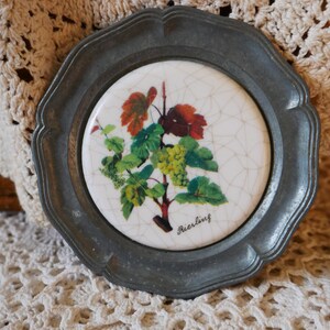 May include: A pewter coaster with a white ceramic insert featuring a grapevine with green leaves and white grapes. The ceramic insert has a cracked glaze and the text "Riesling" is printed in black.