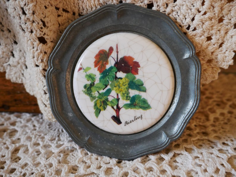 May include: A pewter coaster with a white ceramic insert featuring a grapevine with green leaves and white grapes. The ceramic insert has a cracked glaze and the text "Riesling" is printed in black.