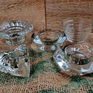 May include: A collection of five clear glass candle holders in various shapes. The holders include a pedestal design, a round design, a small bowl, a faceted design, and a heart-shaped design. The candle holders are displayed on a burlap surface.