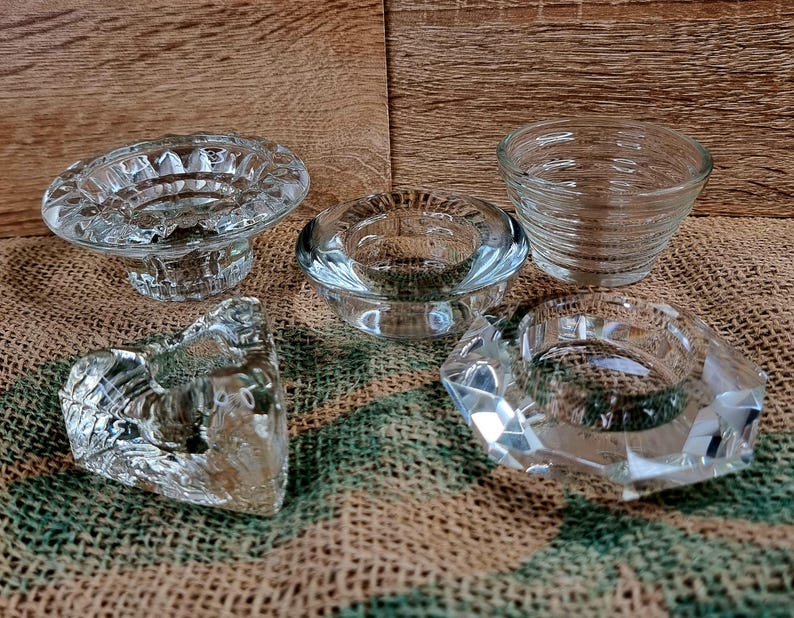 May include: A collection of five clear glass candle holders in various shapes. The holders include a pedestal design, a round design, a small bowl, a faceted design, and a heart-shaped design. The candle holders are displayed on a burlap surface.