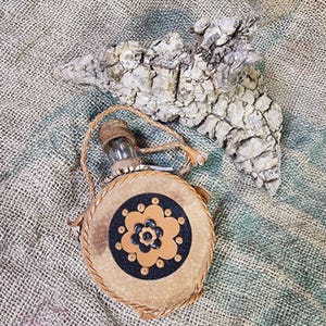 May include: A circular leather flask with a floral design in the centre. It features a glass bottle, cork stopper, and a braided leather strap. A piece of textured, grey bark is positioned behind the flask.