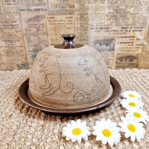 May include: A brown ceramic cheese dome with a decorative design of two mice and a wedge of cheese. The dome is sitting on a brown woven surface with white daisies.