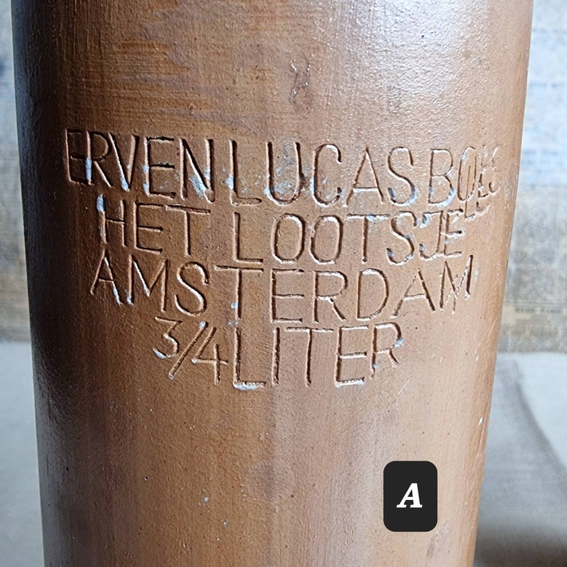 K&ouml;nnte beinhalten: Eine braune Keramikkanne mit wei&szlig;er Beschriftung, die "Erven Lucas Boes Het Lootsje Amsterdam 3/4 Liter" lautet.
