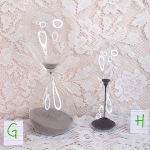 May include: Two clear glass hourglasses with grey sand. The hourglasses are different sizes and sit on a white lace surface. The hourglasses have a classic design, with a bulbous top and bottom connected by a narrow neck.