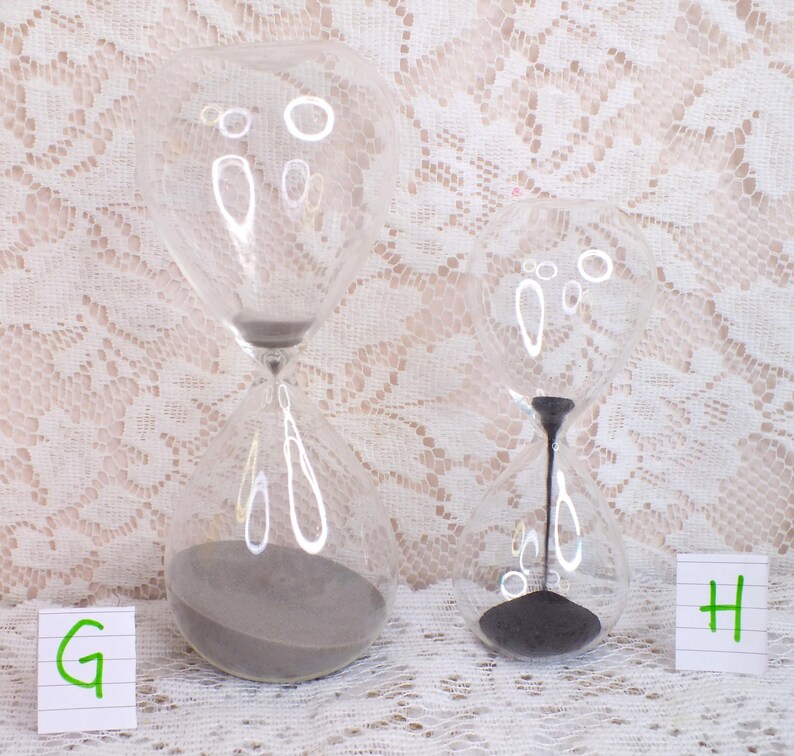 May include: Two clear glass hourglasses with grey sand. The hourglasses are different sizes and sit on a white lace surface. The hourglasses have a classic design, with a bulbous top and bottom connected by a narrow neck.