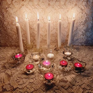 May include: An assortment of lit candles in glass holders. Tall white candles are in ornate holders, while smaller pink candles are in decorative votive holders. The candles are on a lace tablecloth.