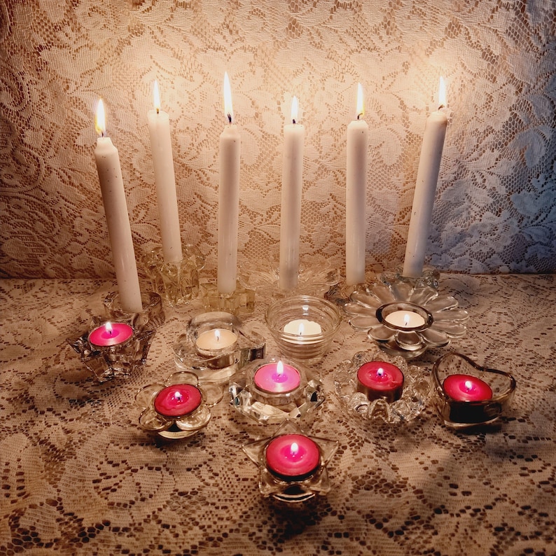 May include: An assortment of lit candles in glass holders. Tall white candles are in ornate holders, while smaller pink candles are in decorative votive holders. The candles are on a lace tablecloth.