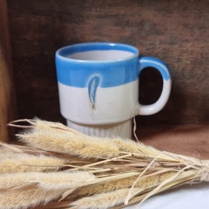 May include: A white ceramic mug with a blue band around the top. The mug has a white glaze with a blue design on the front. The mug is sitting on a wooden surface with dried grass in the foreground.