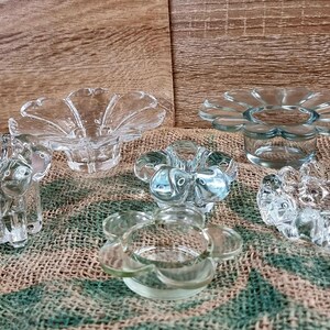 May include: A collection of six clear glass candle holders. The candle holders have a flower-like design with petal-shaped edges. They are arranged on a burlap surface against a wooden background.
