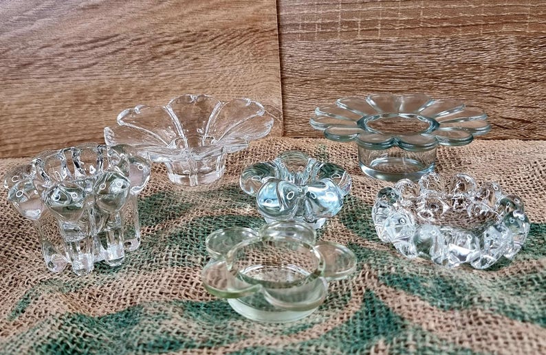 May include: A collection of six clear glass candle holders. The candle holders have a flower-like design with petal-shaped edges. They are arranged on a burlap surface against a wooden background.