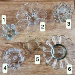 May include: A collection of six clear glass flower-shaped candle holders. Each holder has a unique design, with varying petal shapes and center cup sizes. The holders are arranged on a light brown wooden surface.