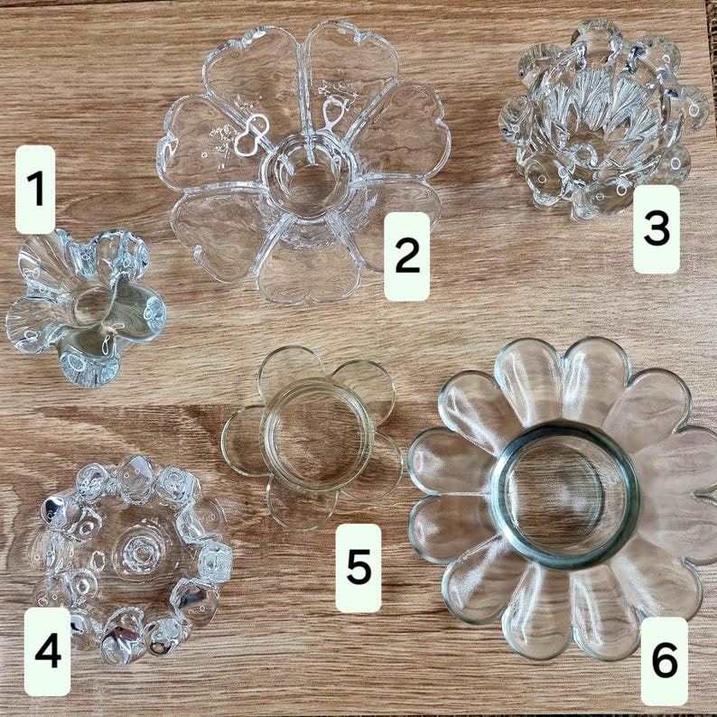May include: A collection of six clear glass flower-shaped candle holders. Each holder has a unique design, with varying petal shapes and center cup sizes. The holders are arranged on a light brown wooden surface.