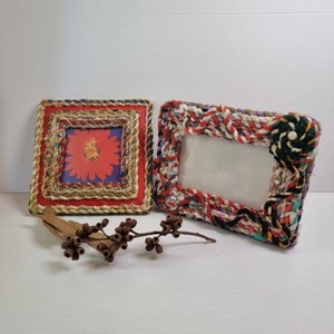 May include: Two handmade picture frames with a rope and fabric design. The frame on the left has a red flower design inside. The frame on the right has a colourful rope design and a black fabric flower.