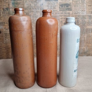 K&ouml;nnte beinhalten: Drei antike Steinzeugflaschen. Zwei sind braun mit einer strukturierten Oberfl&auml;che und eine ist wei&szlig; mit einem blauen Etikett, auf dem "Bohemian Beer" steht.