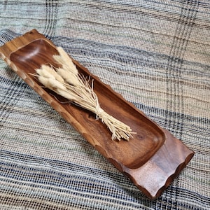 Puede incluir: Una bandeja de servir de madera larga y rectangular con un acabado de madera natural. La bandeja tiene un borde elevado y una superficie lisa. Un manojo de hierbas secas de color marrón claro descansa en la bandeja.