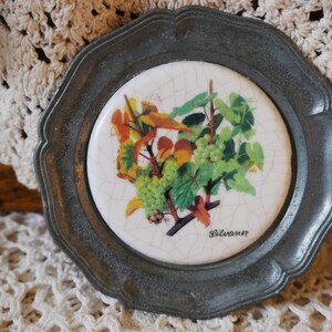 May include: A round, silver-colored coaster with a white ceramic tile inset. The tile features a vintage illustration of a grapevine with green leaves and ripe grapes. The text "Silvaner" is printed in black at the bottom of the tile.