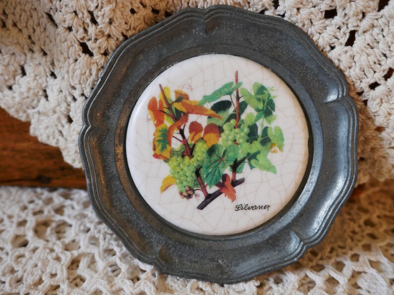 May include: A round, silver-colored coaster with a white ceramic tile inset. The tile features a vintage illustration of a grapevine with green leaves and ripe grapes. The text "Silvaner" is printed in black at the bottom of the tile.