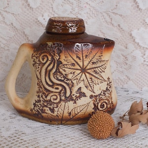 Può includere: Una brocca in ceramica con manico e coperchio, con base beige e parte superiore marrone scuro. La brocca è decorata con motivi a foglie e volute. Un piccolo oggetto rotondo e strutturato e foglie secche sono accanto alla brocca.