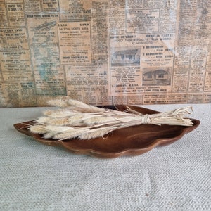 Puede incluir: Un bol de servir de madera con forma de hoja con un manojo de hierbas secas beige en su interior. El bol está sobre una superficie beige texturizada.