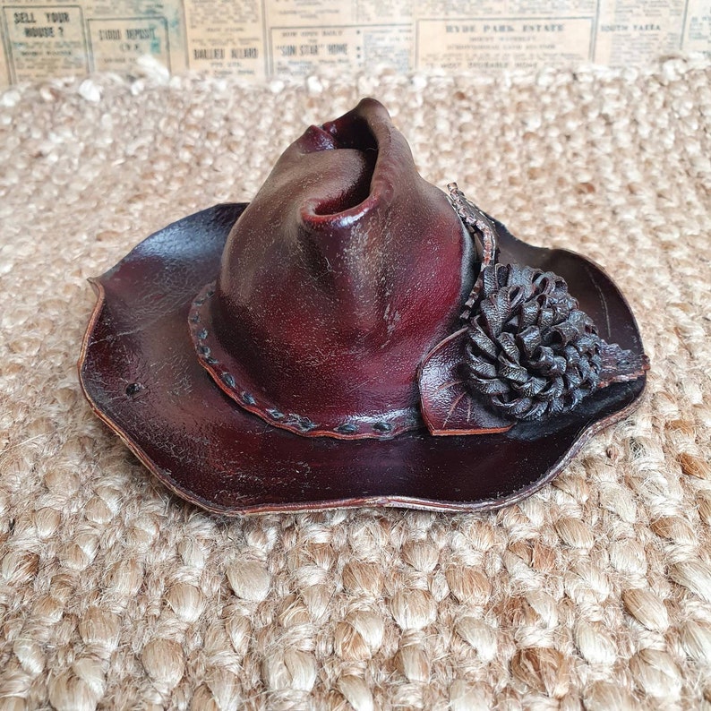 May include: A brown leather cowboy hat with a decorative black leather flower accent. The hat has a wide brim and a pointed crown.