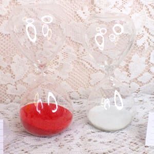 May include: Two clear glass hourglasses with rounded tops and bottoms. One contains red sand, the other white sand. The hourglasses sit on a white lace surface. The image includes two small white cards with green markings.