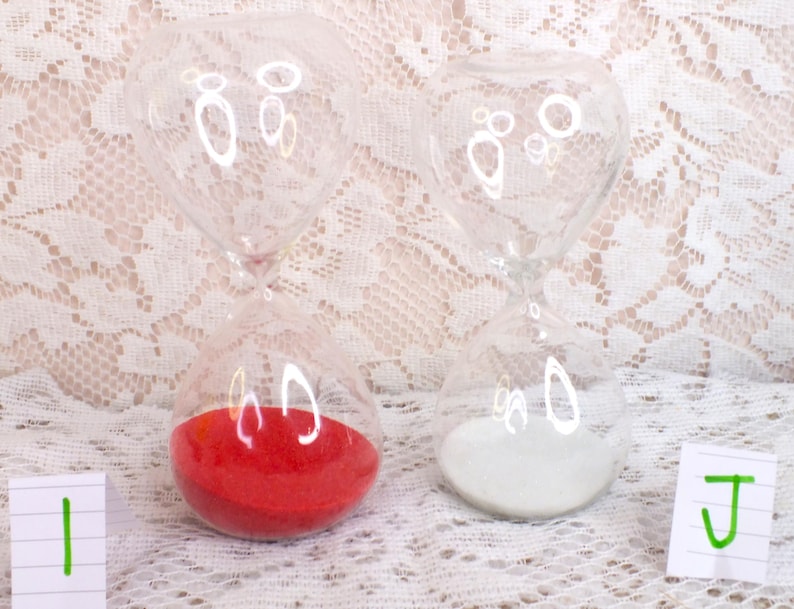 May include: Two clear glass hourglasses with rounded tops and bottoms. One contains red sand, the other white sand. The hourglasses sit on a white lace surface. The image includes two small white cards with green markings.