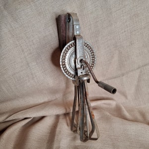 May include: A vintage, silver-coloured, hand-crank egg beater with a wooden handle. The beater has four metal blades and a round gear mechanism.