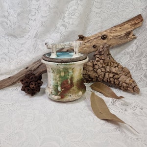 Puede incluir: Un tarro de cerámica con una tapa de diseño único. El tarro tiene una base blanca con detalles marrones, verdes y azules. La tapa tiene un asa blanca y un interior azul.