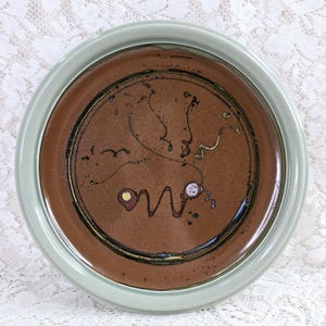 May include: A round ceramic plate featuring a wide, light sage green rim and a dark brown centre. The brown surface is decorated with a thin, dark circular line, abstract black squiggly lines, and two small, circular accents in yellow and white.