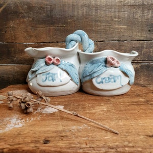 May include: White ceramic double jar with a blue rope handle. The jars are decorated with blue leaves and pink flowers. One jar says "Jam" and the other says "Cream".