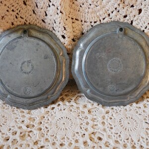 May include: Two silver-toned metal plates with scalloped edges. The back of each plate has a circular design with the text "Atelier d'Art" and "Petites Maisons" in a circle.